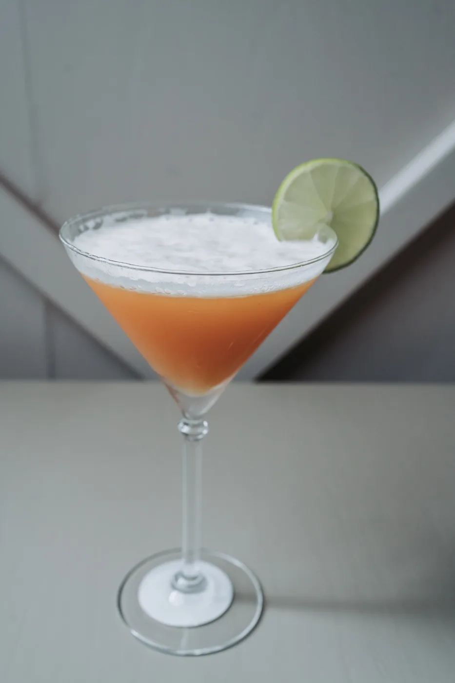 An orange cocktail in a coupe glass garnished with a slice of lime
