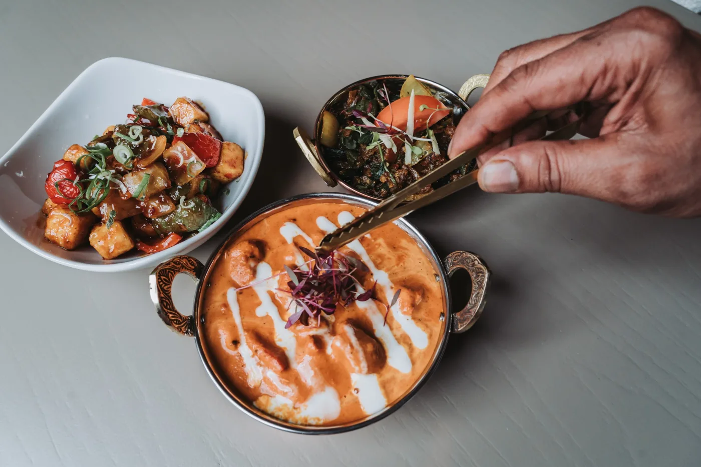 A hand using tongs to garnish a creamy curry in a copper dish, beside bowls of paneer and salad