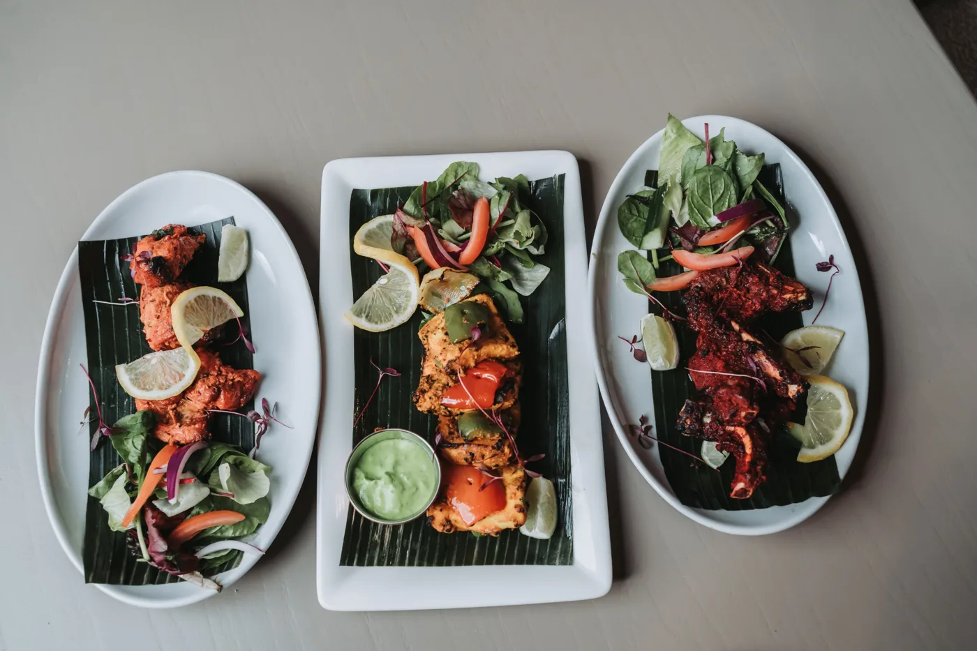 Three plates of tandoori dishes on banana leaves with salad, lemon wedges and a green chutney, seen from above
