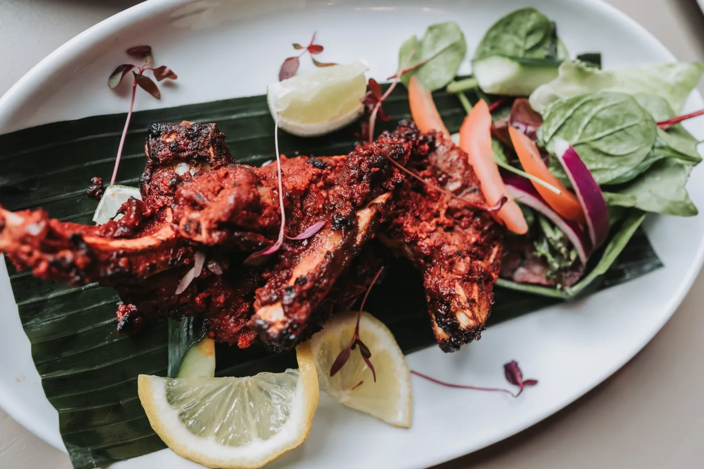 Charred red tandoori pieces on a banana leaf with lemon wedges, red onion and salad leaves on a white plate