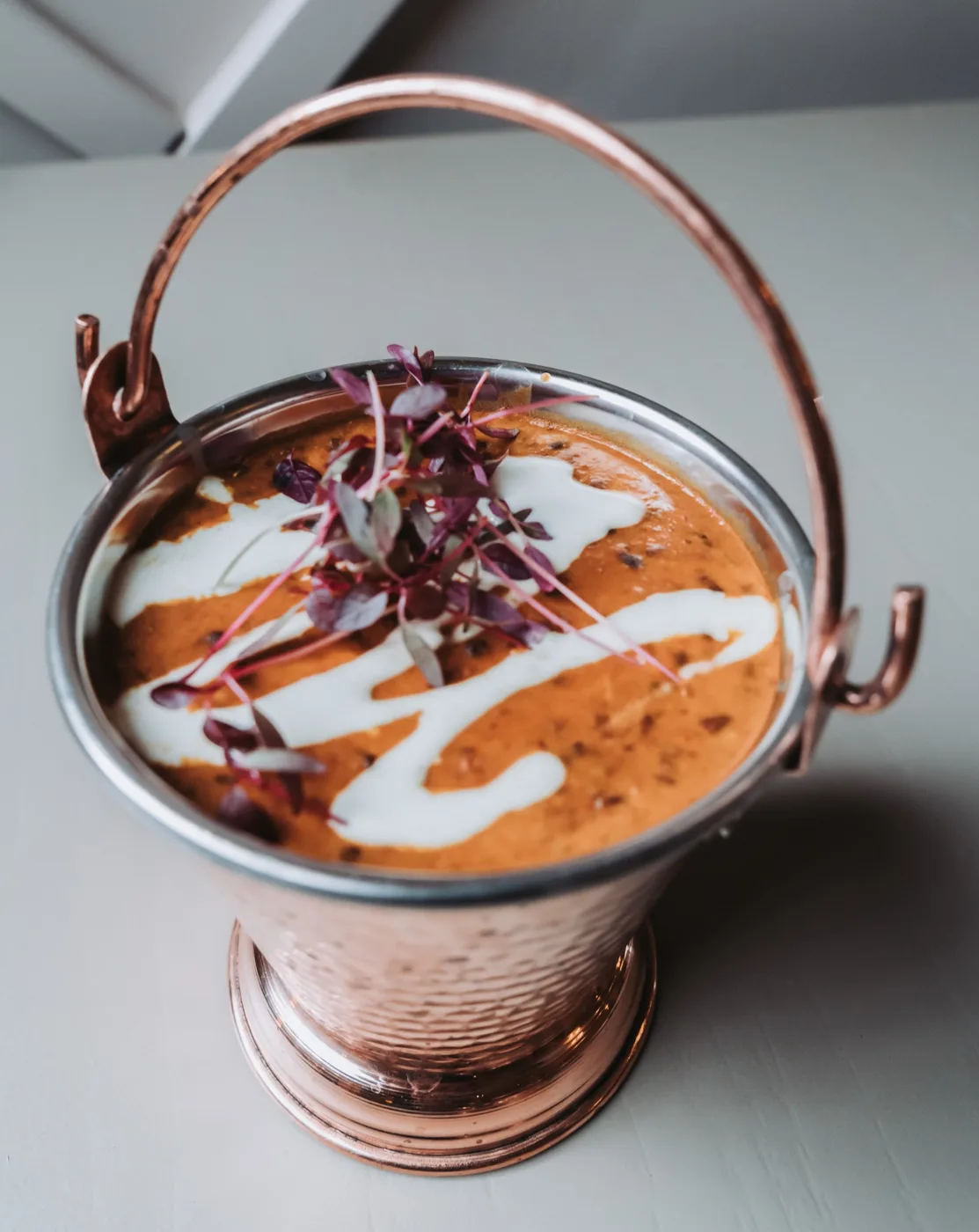 An orange curry swirled with cream and topped with micro herbs, served in a hammered copper bucket with a handle