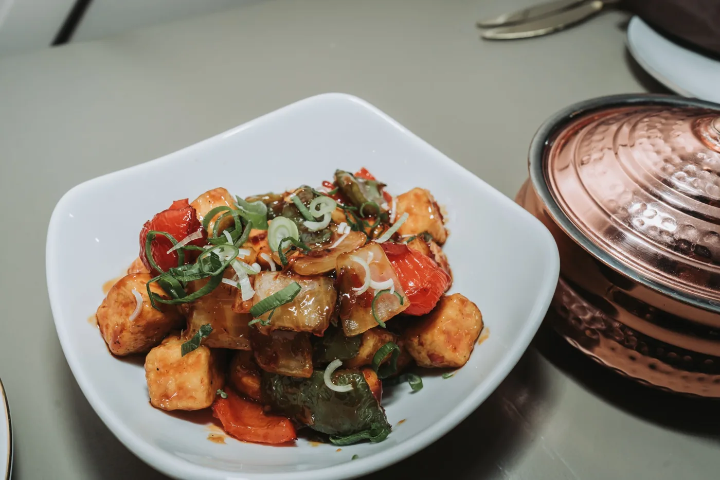 A white bowl of paneer cubes with red and green peppers and spring onion, beside a copper serving pot