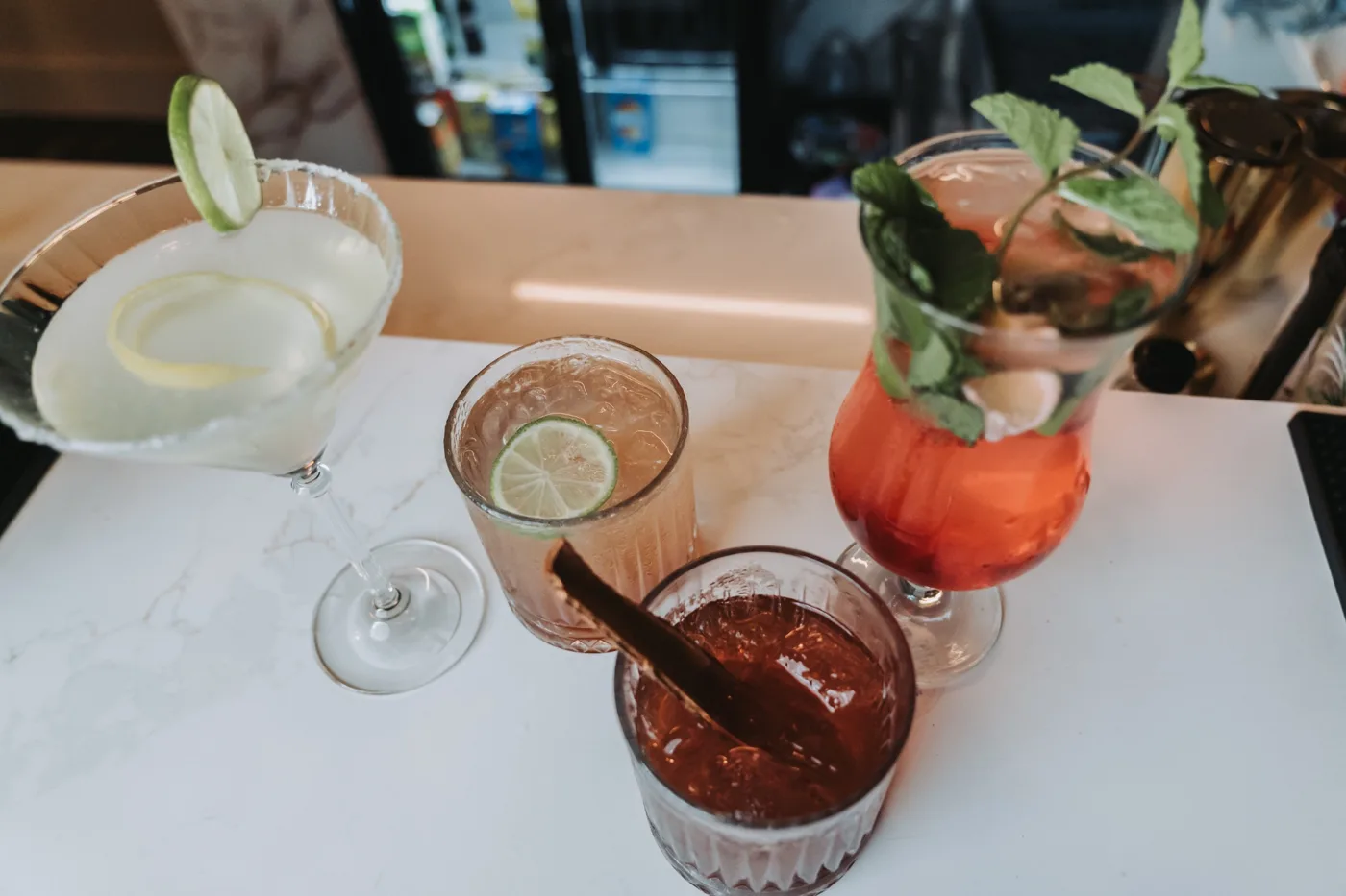 Four drinks on a marble counter, including a lime-garnished cocktail, a tall red drink with mint and a dark drink with a cinnamon stick