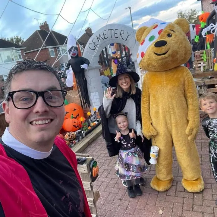 A selfie in front of a Halloween display with a cemetery arch, pumpkins and a Pudsey Bear costume, with a woman and two children in Halloween costumes