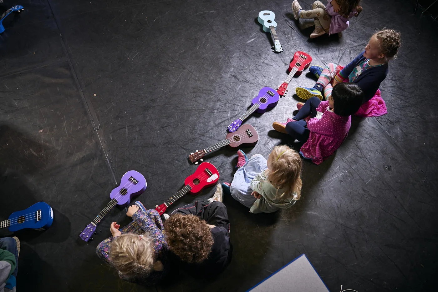 Children seen from above sitting on a black floor among ukuleles laid in a curving line