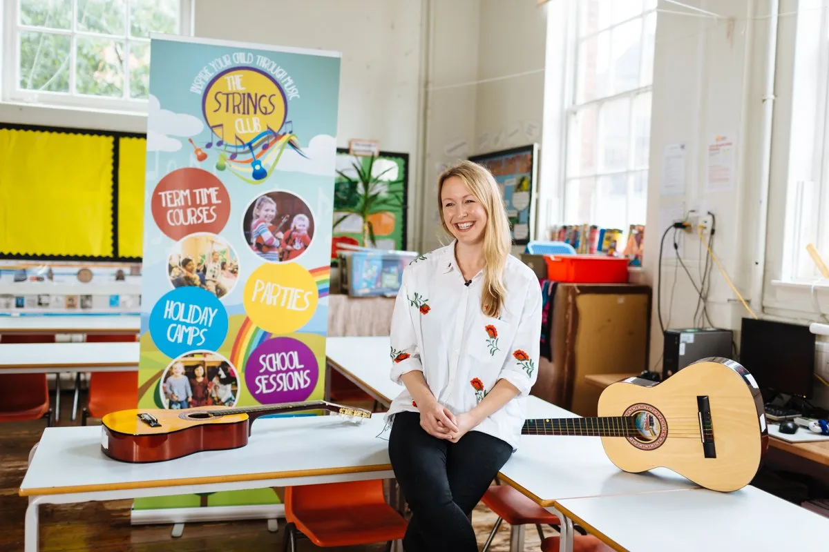 Amy Cunningham sitting on a table in a classroom between two acoustic guitars, beside a Strings Club pull-up banner listing team time courses, parties, holiday camps and school sessions
