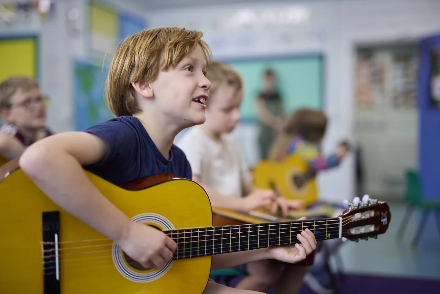 A boy playing an acoustic guitar in a classroom, with other children behind him