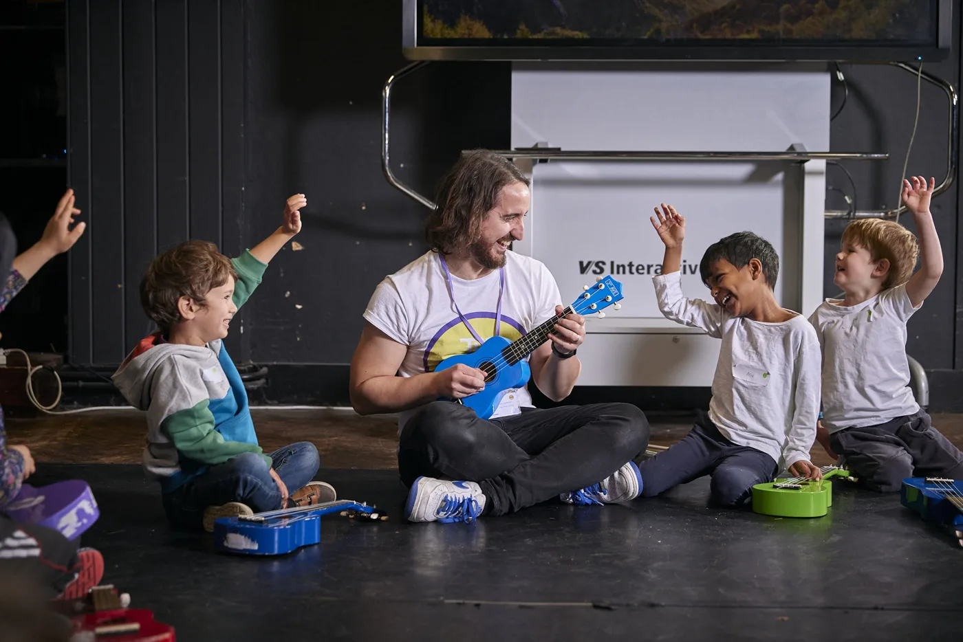 A man in a Strings Club T-shirt sitting cross-legged on the floor playing a blue ukulele, with children around him raising their arms