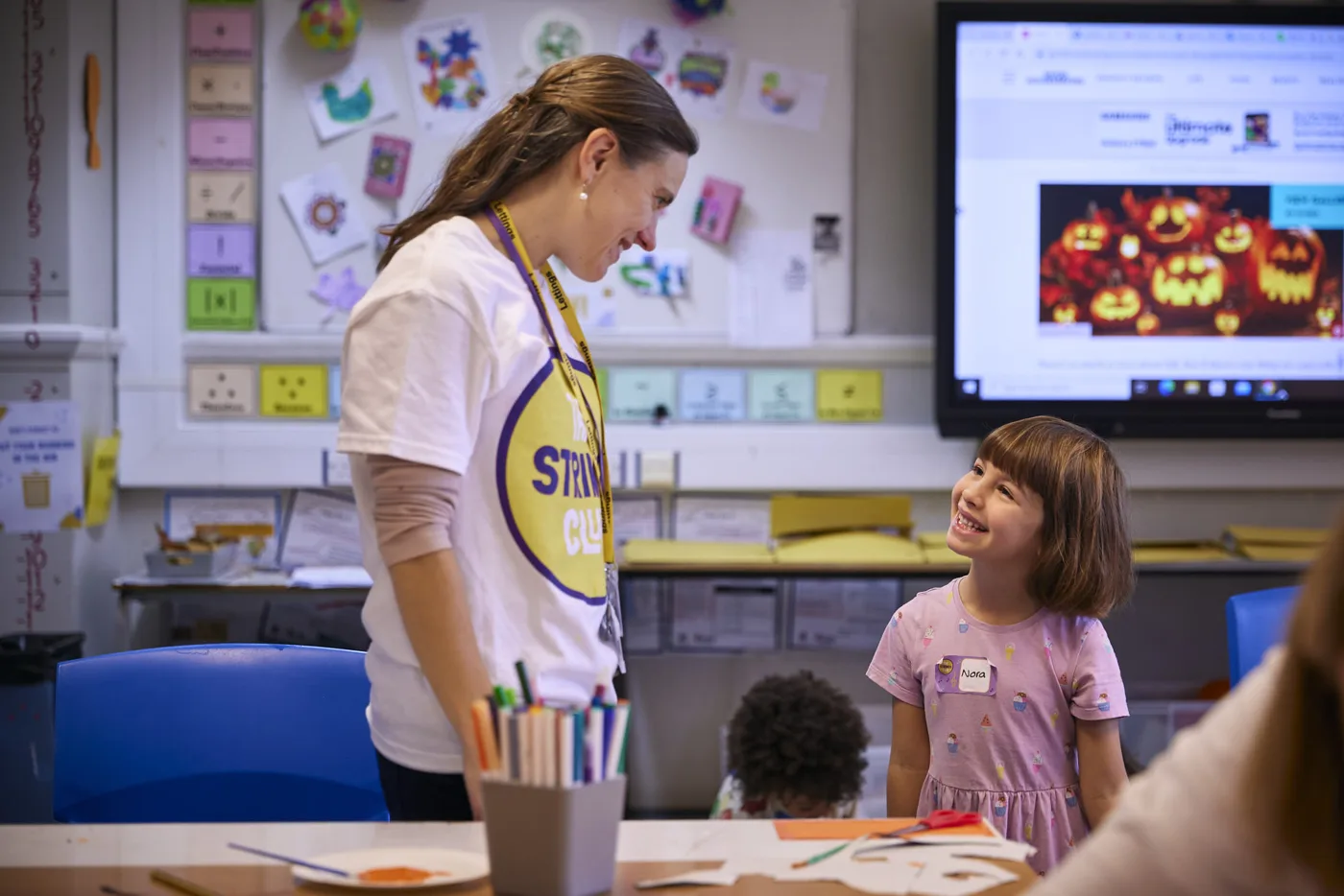 A woman in a Strings Club T-shirt smiling at a girl in a classroom