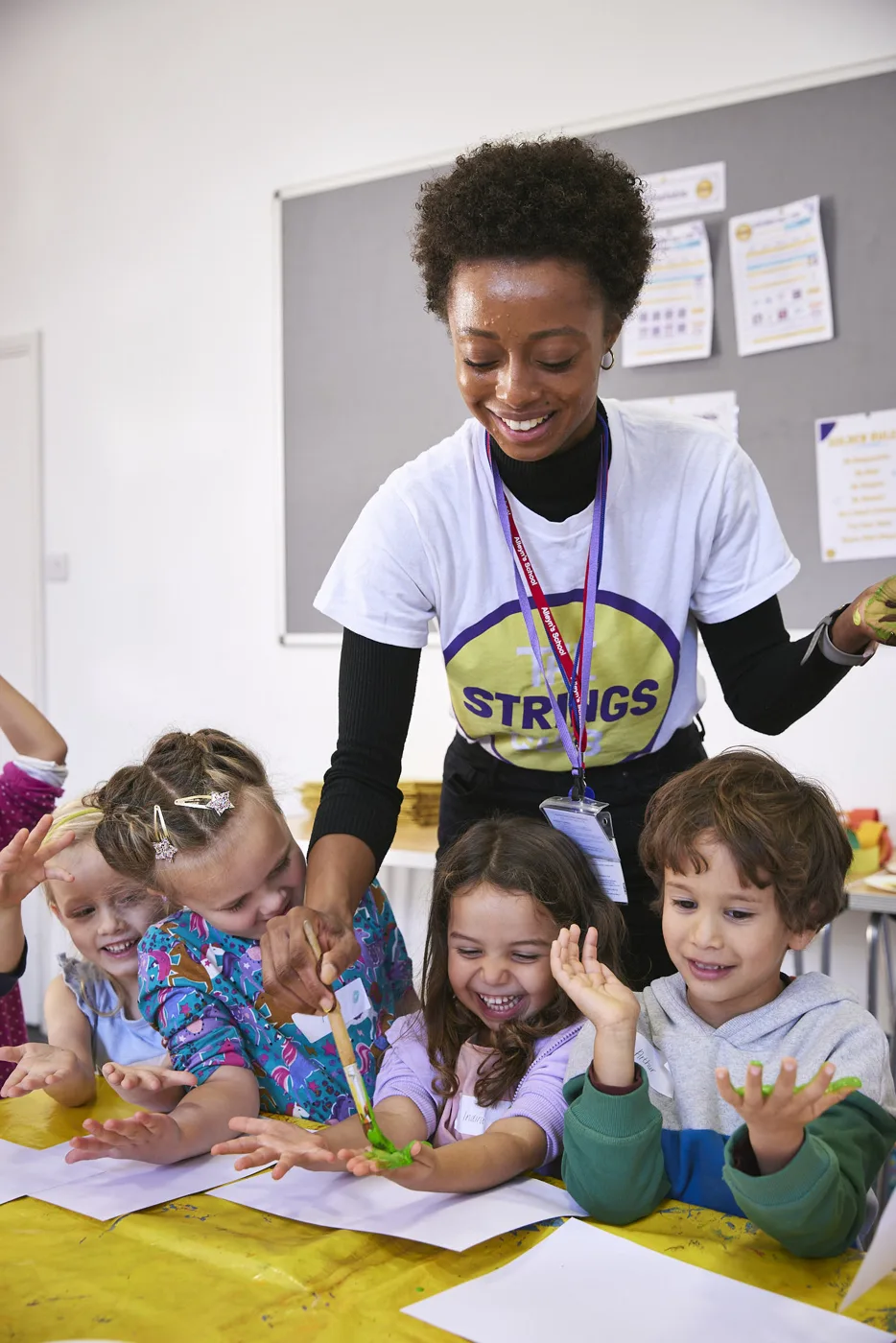 A woman in a Strings Club T-shirt leaning over a table of young children holding up their hands, with paper and craft materials on the table