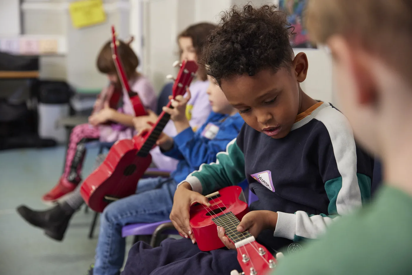A boy looking down at a red ukulele, sitting in a row of children holding ukuleles