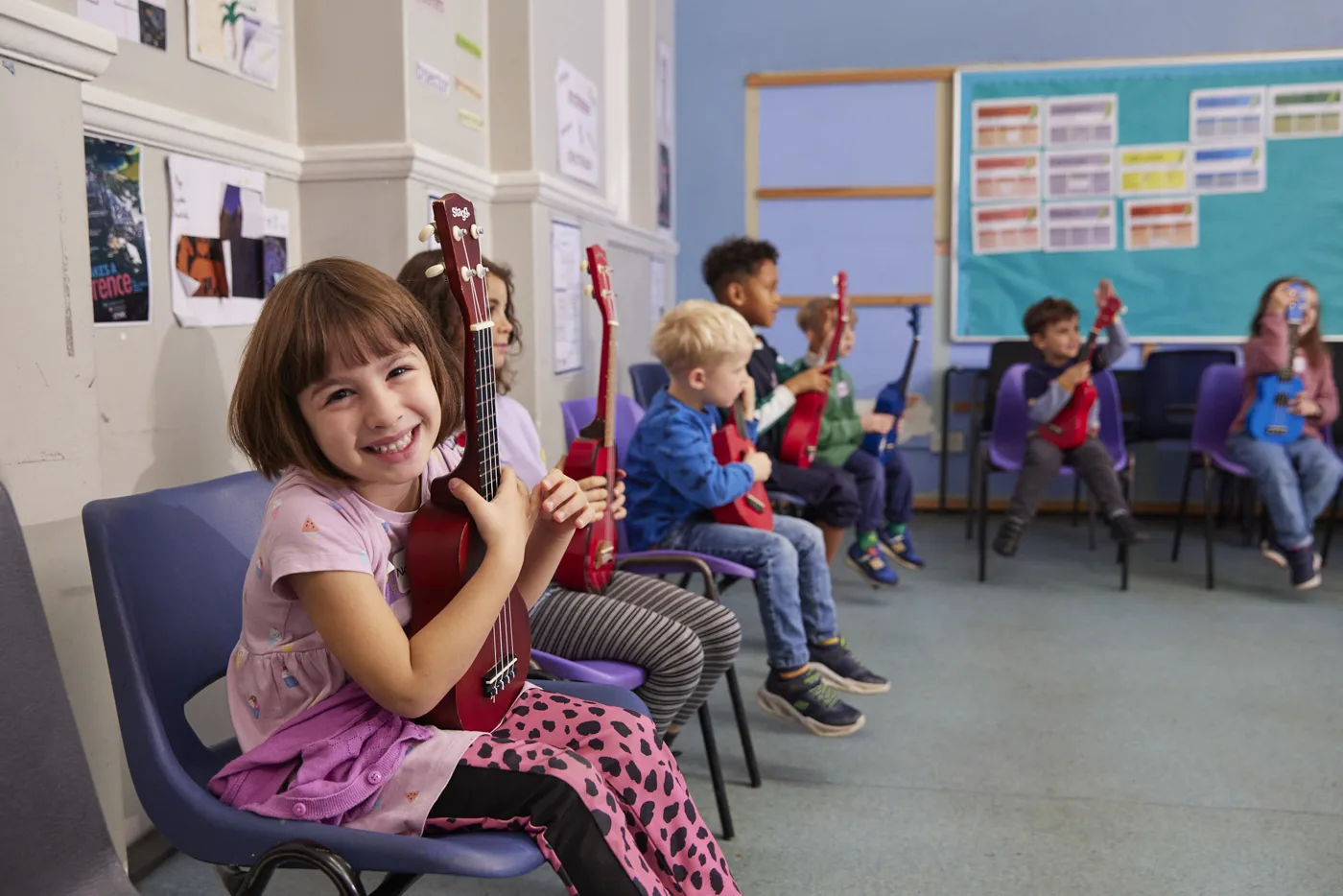 A girl holding a ukulele at a Strings Club holiday camp, with other children playing behind her